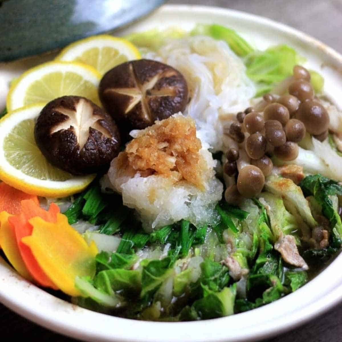 a bowl of japanese hot pot with lemon  slices, noodles, cabbage, carrots and mushrooms