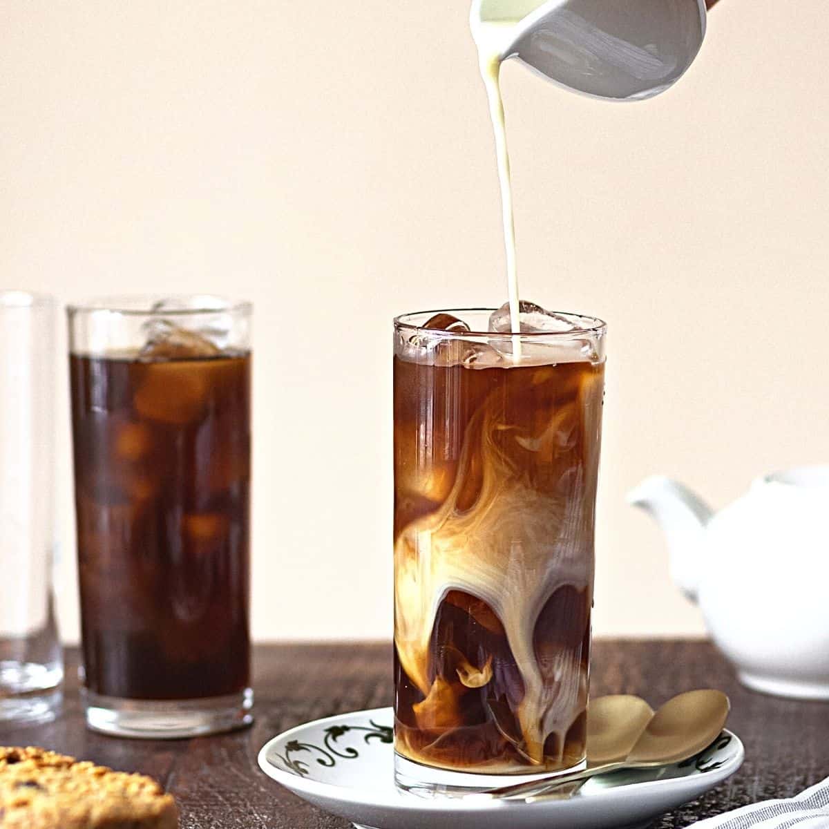 cream being poured into a glass with coffee and ice with another glass of iced coffee near